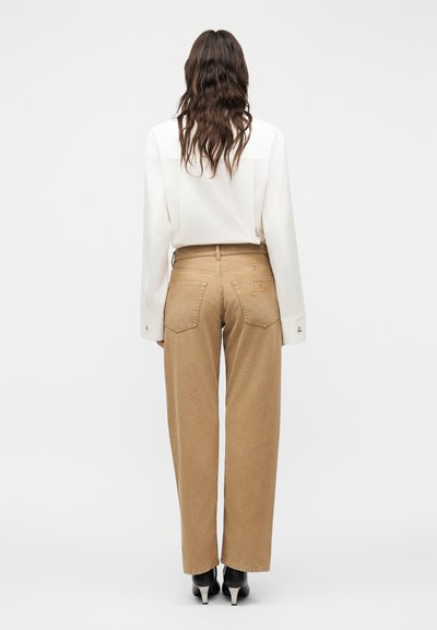 Γυναίκα με μακριά κυματιστά καστανά μαλλιά, που φοράει λευκό πουκάμισο με μακριά μανίκια, μπεζ παντελόνι ψηλόμεσο και μαύρα μποτάκια με τακούνι, που στέκεται με την πλάτη.