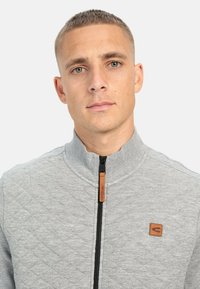 Veste zippée gris clair avec un design matelassé, col haut, fermeture éclair noire et petite étiquette en cuir marron sur la poitrine gauche.
