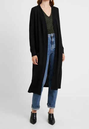 Kvinde iført lang sort cardigan med åben front, olivengrøn hækletrøje, blå opsmøgede jeans og sorte hælede mules, stående mod en hvid baggrund.