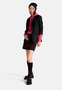 Černý blejzr přes růžovou mikinu s grafickým potiskem, hnědá mini sukně, černé podkolenky a černé sandály se popruhy.