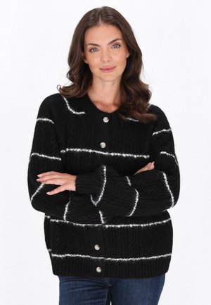 Frau mit welligem braunem Haar, die einen schwarzen, geknöpften Pullover mit weißen horizontalen Streifen trägt, steht mit verschränkten Armen und lächelt sanft.