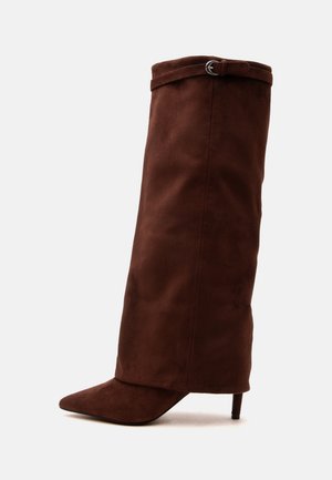 Botte haute en suède marron foncé, avec un bout pointu, un talon haut et fin, et une petite lanière avec boucle près du haut.