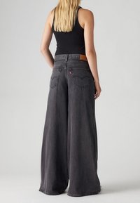 Szerokie czarne dżinsy z wysokim stanem, z brązową skórzaną łatą z tyłu oraz subtelnym haftem na pasku i kieszeniach.