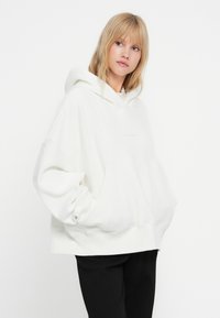 Felpa oversize bianca con cappuccio, dotata di tasca a marsupio e polsini a coste, con un logo minimale in rilievo sul petto.