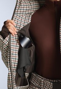 Jakna s uzorkom kariranog zuba nepravilno je otvorena, otkrivajući tamnu unutrašnjost s džepom u kojem se nalazi crni predmet. Jakna ima složenu teksturu i dizajn.