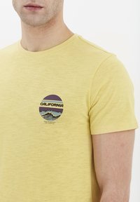 Gul kortærmet t-shirt lavet af blødt bomuldsstof med rund halsudskæring. Har et grafisk tryk med "CALIFORNIA" og bølgedesign på brystet.
