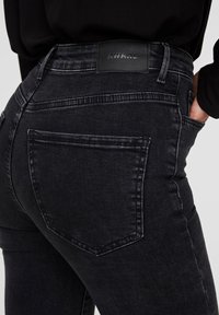 Vaqueros de mezclilla negra con cintura alta, con un parche de logo en la parte trasera y un diseño clásico de cinco bolsillos. Textura suave y forma ajustada.