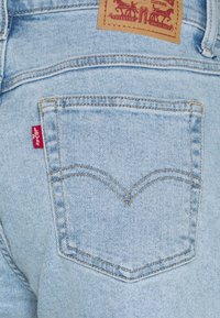 Bolsillo de unos pantalones vaqueros de denim azul claro con un diseño de costura en forma de V, pequeña etiqueta roja en el lateral y un parche de cuero marrón en la parte superior.