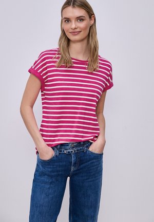 Kobieta z rękami w kieszeniach, ubrana w różowo-biały prążkowany krótki rękaw i niebieskie dżinsy na jednolitym jasnym tle.