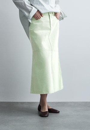 Vrouw met een lichtgroene midi denimrok met voornaad, wit overhemd met lange mouwen, handen in de zakken en donkerbruine platte schoenen, staand op een grijze vloer.