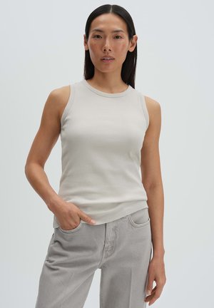 Asiatische Frau mit langen schwarzen Haaren, die ein ärmelloses, beiges, geripptes Oberteil und hellgraue Jeans trägt, steht mit einer Hand in der Tasche.