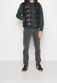 Schwarze Steppweste mit Kapuze über einem grünen Sweatshirt, kombiniert mit grauen Jeans und schwarzen Schuhen mit Gummisohlen. Einfache Nahtdetails sichtbar.