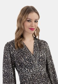 Leopardmønstret kjole i blødt stof, med dyb V-hals, lange puffede ærmer og drapering ved taljen. Farveskalaen inkluderer sort og beige.