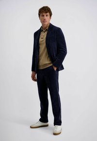 Man in een marineblauw corduroy pak met een beige polo shirt en witte sneakers, staande tegen een effen lichte achtergrond.