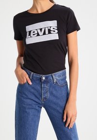Černé bavlněné tričko s bílým logem "Levi's" v obdélníkovém designu, spárované s vysokými modrými džíny z denimové látky s vybledlým vzhledem.