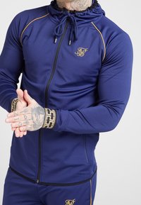 Navy athletic zip-up jacket with gold accents, a drawstring hood, and patterned cuffs. Features a fitted design and smooth texture.