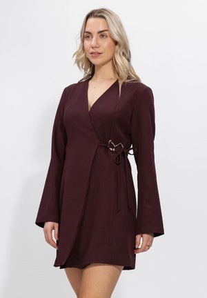 Blonde vrouw die een lange mouw, diep bordeauxrode wikkeljurk draagt met een zijstrik en zilveren gespsluiting.