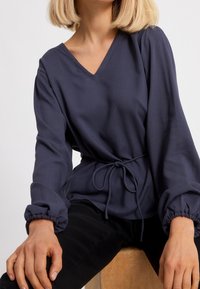 Mörkblå V-ringad blus med samlade manschetter och knytning i midjan; slätt tyg med avslappnad passform. Svarta byxor delvis synliga.