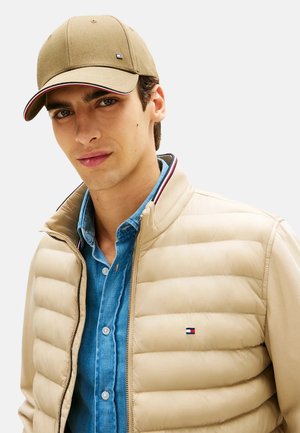 Jeune homme portant une veste matelassée beige, une chemise bleu boutonnée et une casquette de baseball beige assortie avec un petit logo sur les deux vêtements.