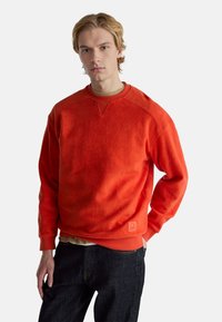 Červená mikina z měkkého fleecu, s kulatým výstřihem a žebrovanými manžetami. Na levé dolní straně je malá logo.