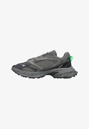 Zapatilla deportiva gris con suela texturizada, parte superior de malla y material sintético, y una pequeña lengüeta de tirador en color verde brillante en el talón.