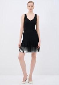 Abito nero a coste con scollatura a V e orlo plissettato, che si trasforma in un motivo più chiaro nella parte inferiore. Modella con tacchi bianchi.