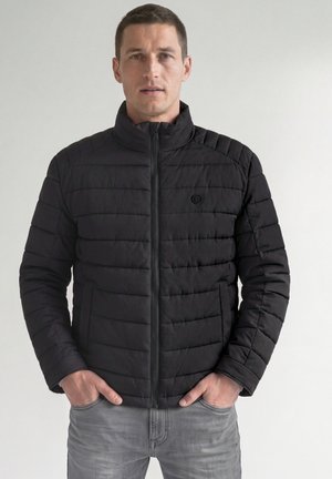 Mann, der eine schwarze gesteppte Jacke mit hochstehendem Kragen trägt und die Hände in den Taschen hat, steht vor einem schlichten hellen Hintergrund.