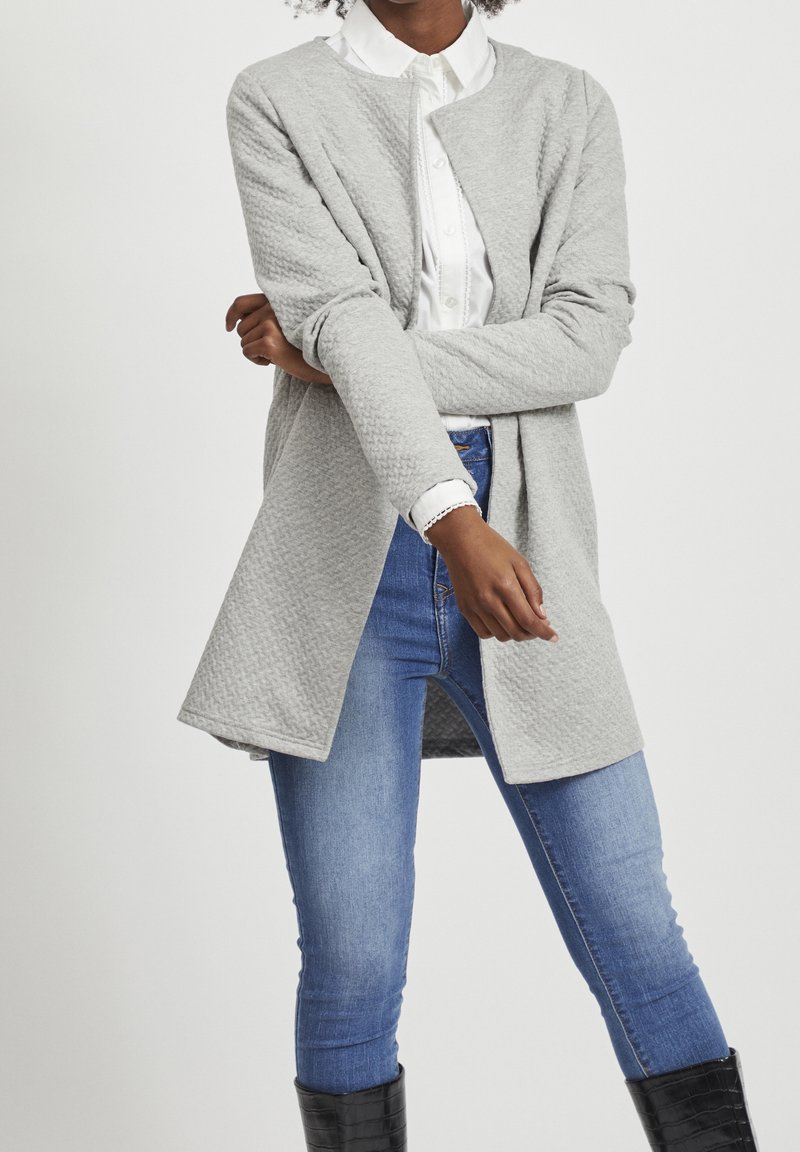 Grijze textuur lange cardigan met open voorkant, gedragen over een witte blouse met kraag en blauwe spijkerbroek; heeft lange mouwen en een ontspannen pasvorm.