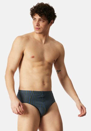 Joven con cabello rizado vistiendo calzoncillos azules con rayas verticales amarillas y grises, posando contra un fondo liso claro.