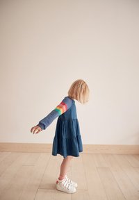 Vestito in denim con design a strati, parte superiore blu con maniche a strisce arcobaleno. Sneakers bianche con strisce, pavimento in legno chiaro, sfondo semplice.