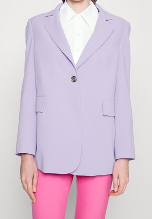 Personne portant un blazer violet clair avec deux poches avant et un seul bouton sur une chemise blanche à col et un pantalon rose vif.