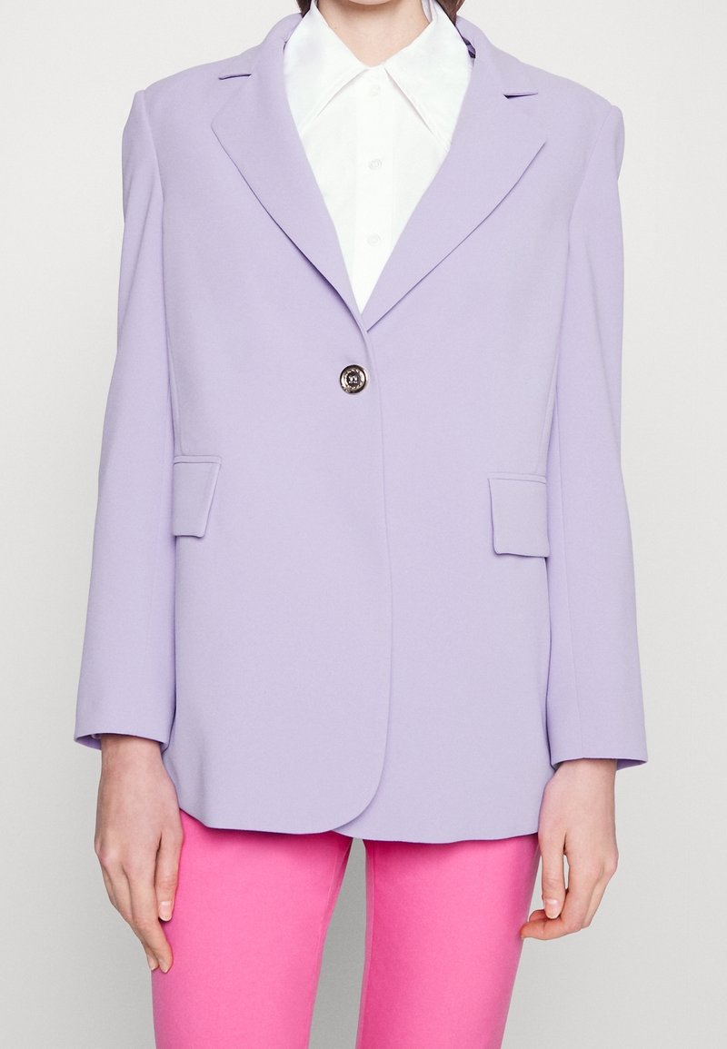 Personne portant un blazer violet clair avec deux poches avant et un seul bouton sur une chemise blanche à col et un pantalon rose vif.