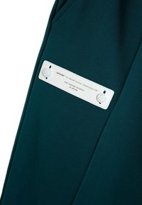 Un bolsillo de tela verde oscuro con una etiqueta blanca. La etiqueta presenta texto en relieve y dos botones a presión, lo que proporciona detalles de marca y acentos de diseño.