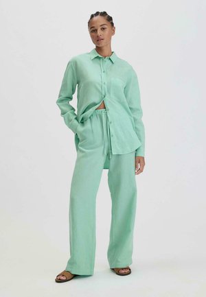 Lichtgroene linnen shirt met knoopsluiting en zak, gecombineerd met bijpassende loszittende broek. Model draagt bruine sandalen.