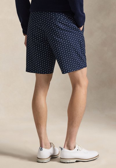 Muž oblečený v tmavě modrých šortkách s malými bílými květinovými vzory, tmavě modrém svetru a bílých sportovních botách, stojící s rukama v kapsách.