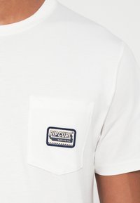 Tricou alb din bumbac cu guler rotund și o buzunar pe pieptul stâng, având un patch cu logo-ul "Rip Curl" în nuanțe de albastru și crem.