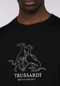 Černý pletený svetr s bílou vyšívanou grafikou dvou psů. Pod designem je bílé textové logo "TRUSSARDI GENTLE SOCIETY".