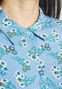 Camicia floreale blu con pois bianchi, colletto e bottoni azzurri chiari. Il motivo floreale include fiori arancioni e blu con steli verdi.