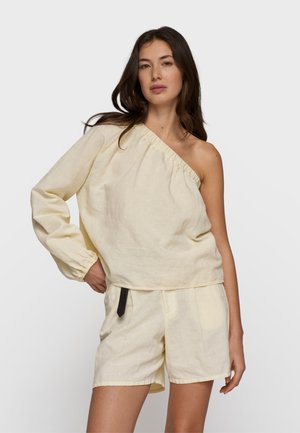 Kobieta w kremowej, jednokołnierzowej bluzce z długim rękawem, połączonej z pasującymi szortami, stojąca z ręką na biodrze na jednolitym tle.