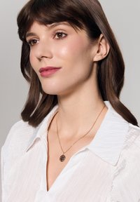 Złoty naszyjnik z okrągłym wisiorkiem przedstawiającym wzór drzewa, noszony na białej, fakturowanej koszuli, podkreślający misterny detal wisiorka.