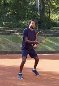 Tennis spiller i mørk blå treningstøy, holder en racket med oransje detaljer, på en leirbane omgitt av grøntanlegg og steintrapper.