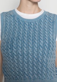 Pull sans manches en tricot bleu avec un motif en torsades et une finition texturée. Encolure ronde et teintes plus claires contrastantes.