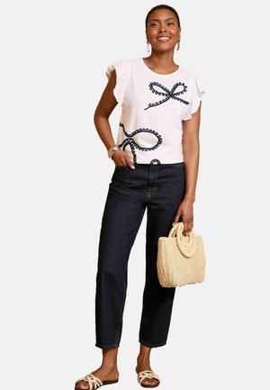 White short-sleeve top with black loop design, paired with dark denim pants, and a woven straw handbag. Flat sandals complete the outfit.