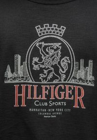 Μαύρο μπλουζάκι με γραφικό λευκού λιονταριού μέσα σε κυκλικό πλαίσιο, στοιχεία ορίζοντα και κόκκινο κείμενο "HILFIGER CLUB SPORTS".