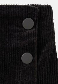 Čierny kordový materiál s vertikálnymi pruhmi, dvoje čierne okrúhle gombíky ako výrazný detail blízko horného okraja.