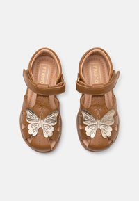 Brązowe skórzane sandały z wyciętym wzorem, regulowanym paskiem i dekoracyjnym metalowym motylem na przodzie.