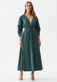 Teal pitkä mekko syvällä V-kauluksella, vyötäröltä kapeaksi taitaen ja pussimaisilla hihoilla. Sujuva pinta, lattianpituinen, yhdistettynä nude-värisiin hihnasandaaleihin.