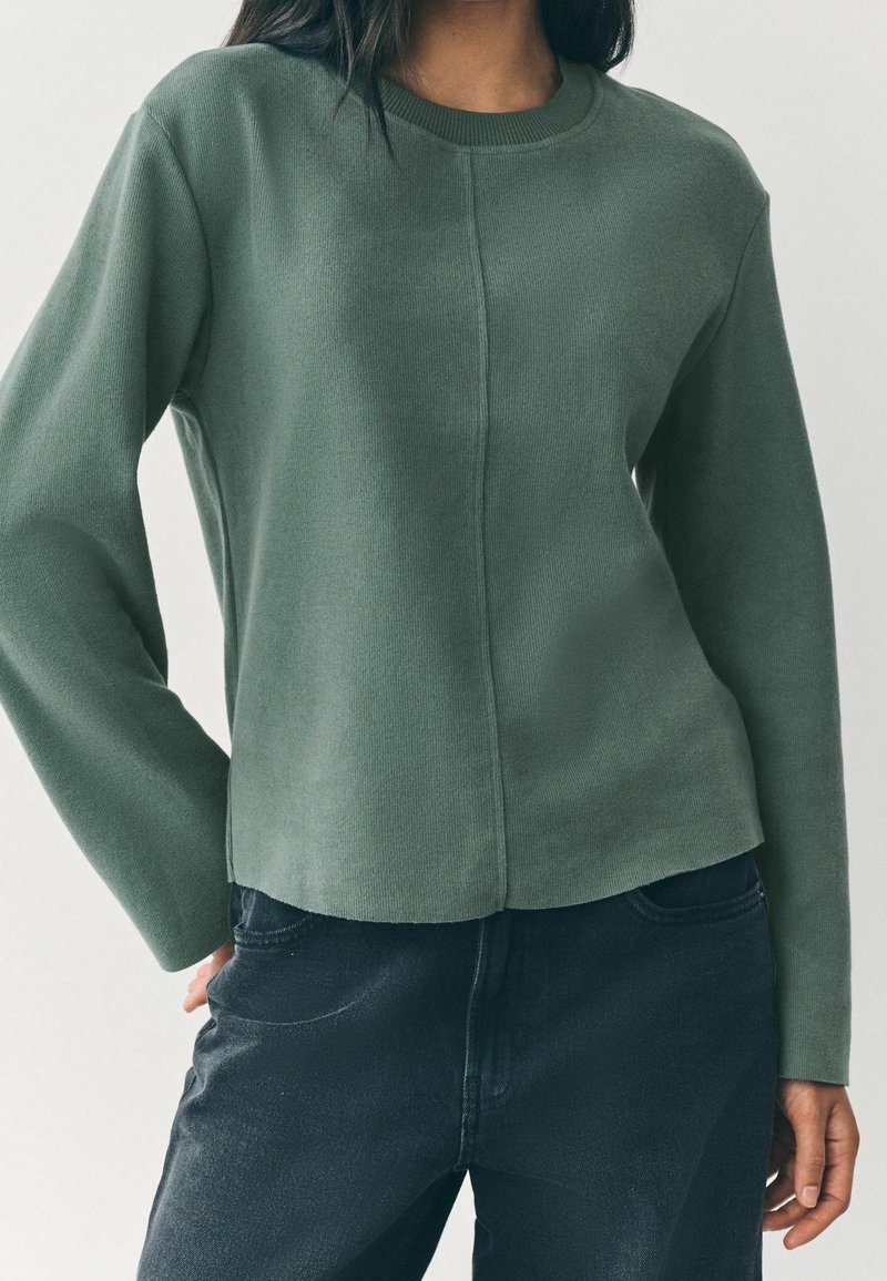 Femme portant un haut en maille vert olive à manches longues associé à un jean bleu foncé, debout avec une main dans la poche devant un fond uni.