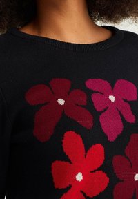 Schwarzer Strickpullover mit großen Blumenmustern in Rot- und Rosatönen, mit weißen Mittelpunkten und einem gerippten Ausschnitt.