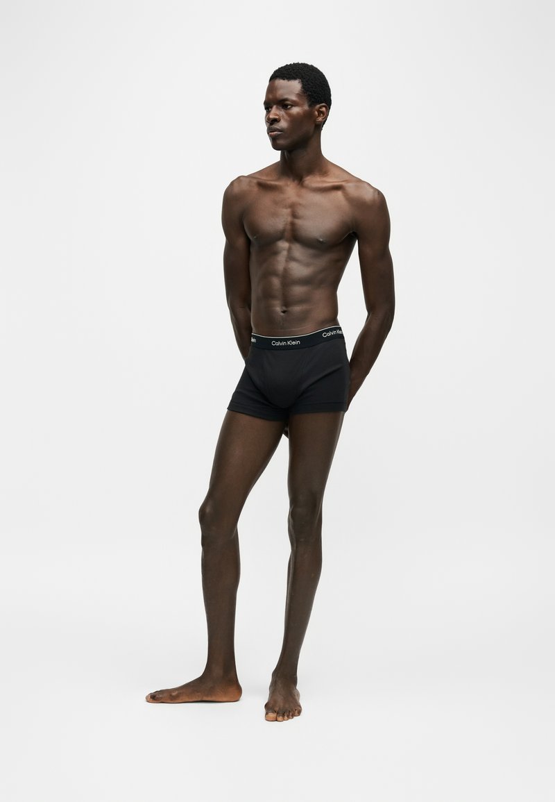 Homme musclé debout pieds nus portant un boxer Calvin Klein noir sur un fond uni gris clair.